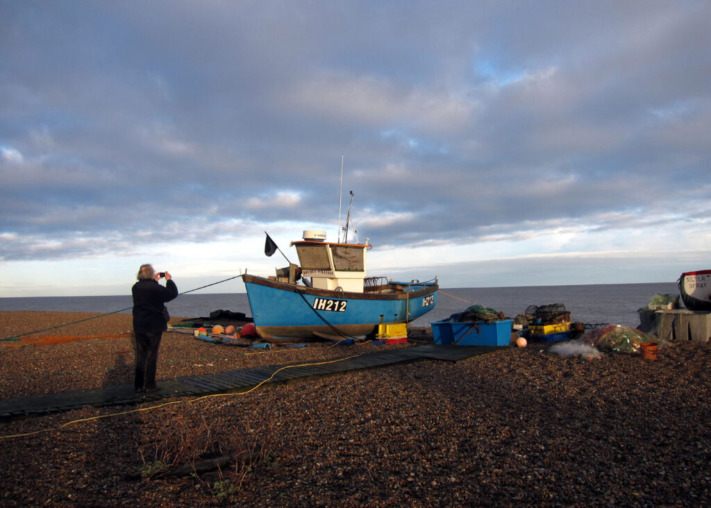 dad photographing a fishing boat with tackle on the shingle beach, sea and blue late afternoon sky with clouds