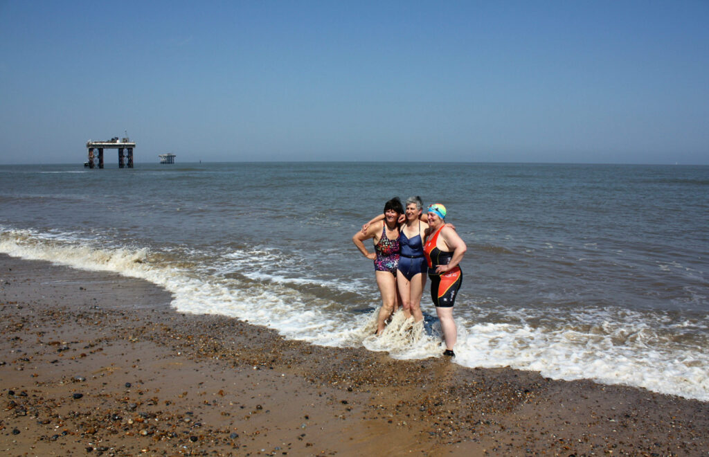 three swimmers with arms round each other at the edge of the sea, blue sky, power station outfall structures
