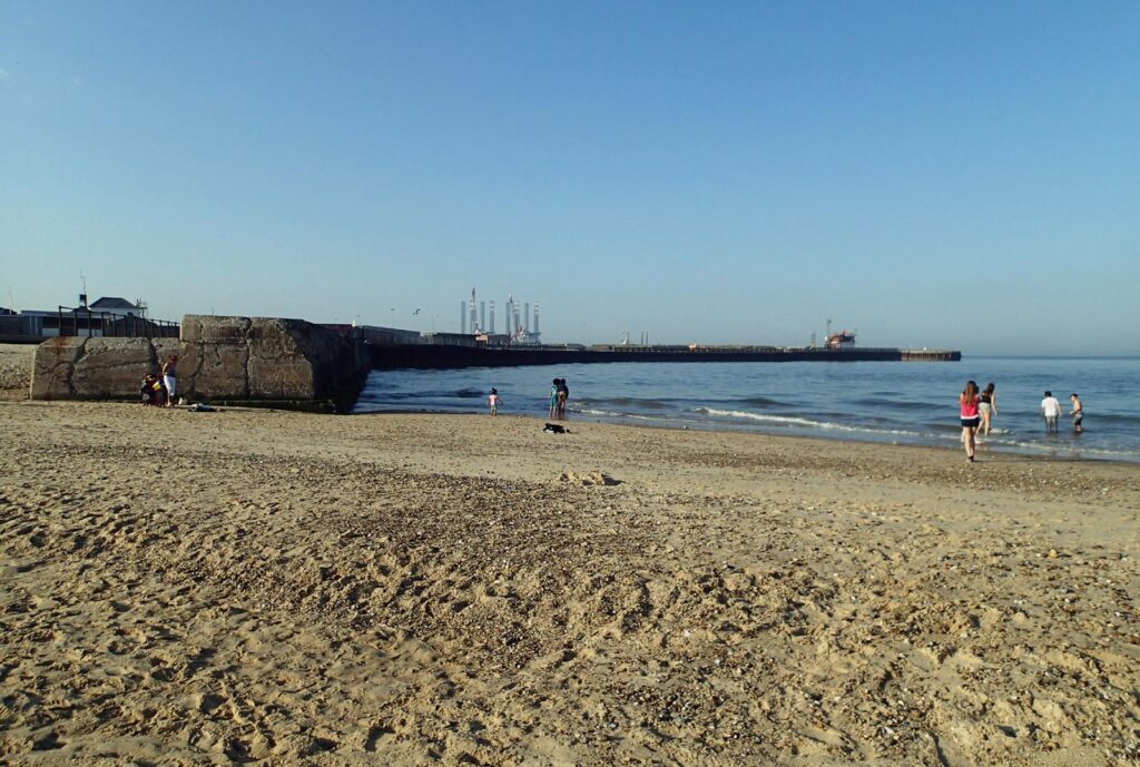Sandy beach, harbour wall and distant towers, people paddling in the sea