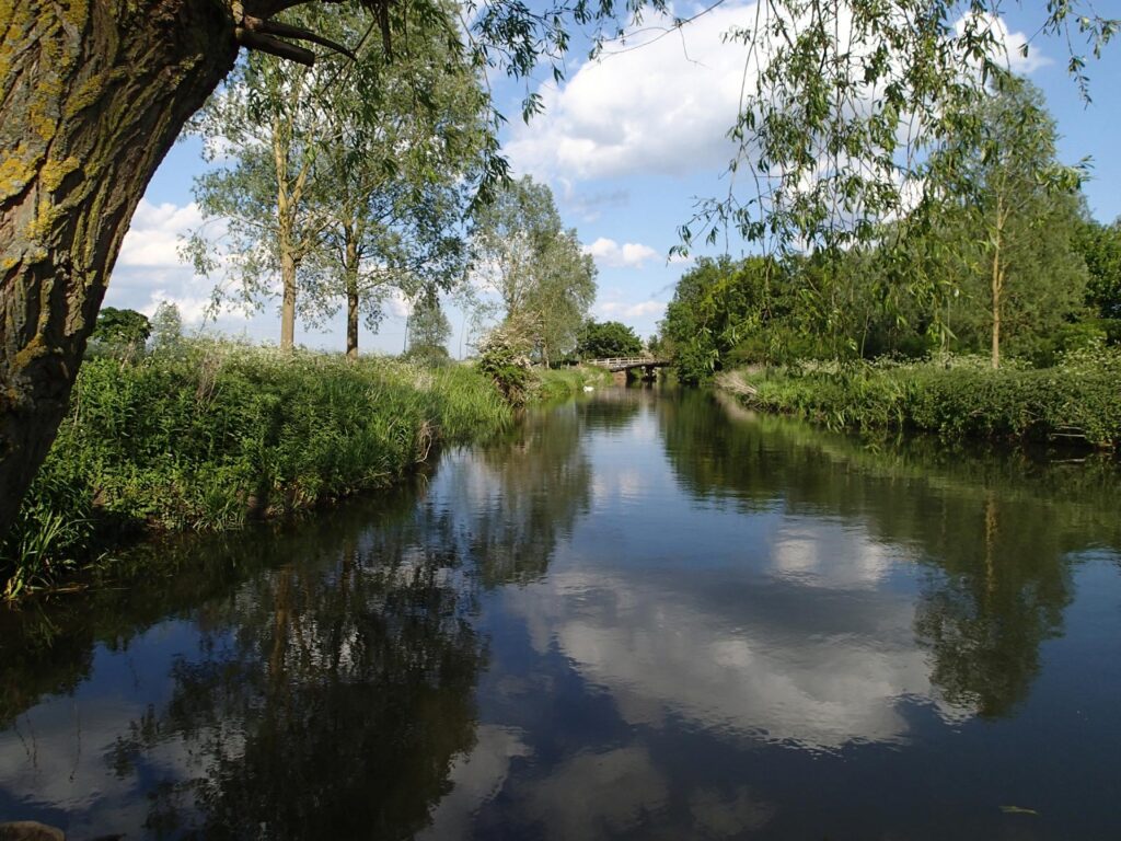 along the river towards a bridge, trees on the left, clouds and blue sky reflected in the water