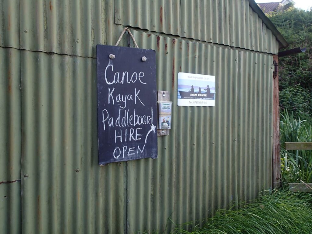corrugated iron shed with signs reading “Canoe Kayak Paddleboard HIRE OPEN, Iken Canoes”