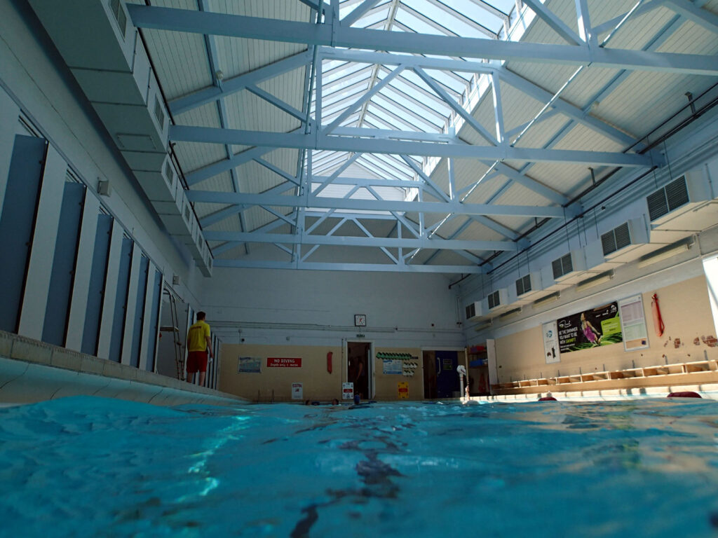 an indoor swimming pool lined with changing rooms and benches, lifeguard in yellow and red, light through atrium above