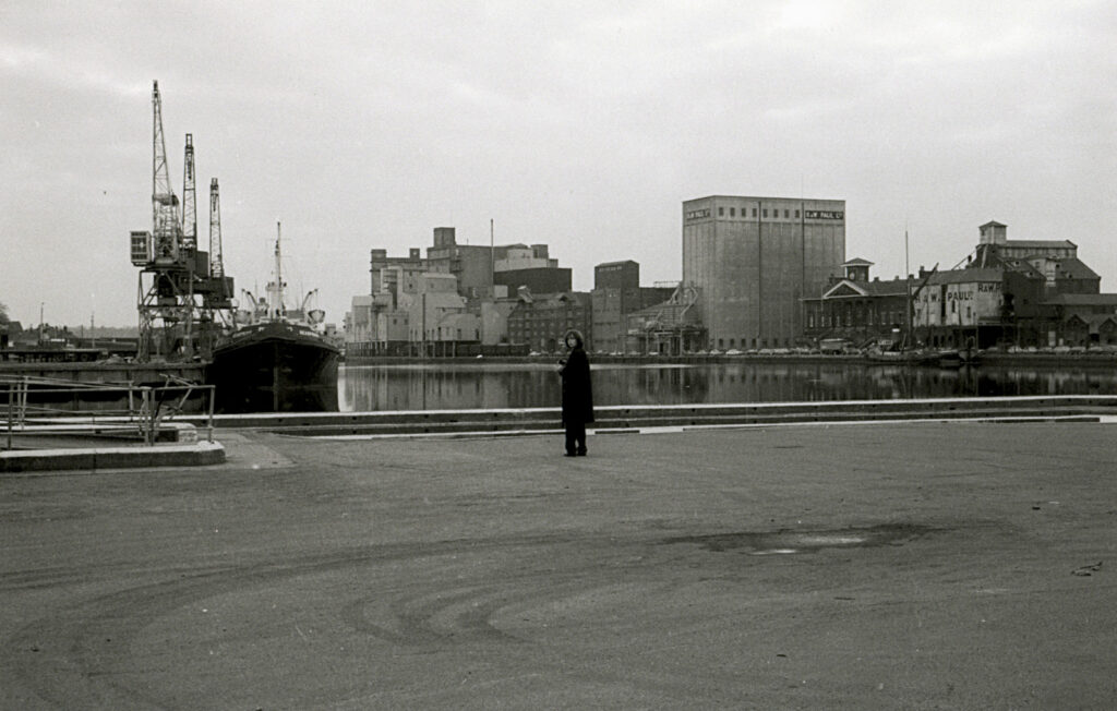 Looking across the docks to buildings and barge, crane and boat, Nick in long black coat looking back at camera, black-and-white photo