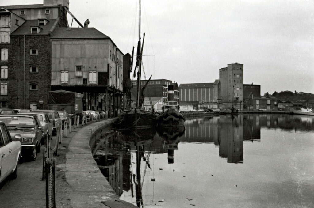 Dockside buildings, one on stilts, sailing barge, water and old-fashioned cars, black-and-white photo