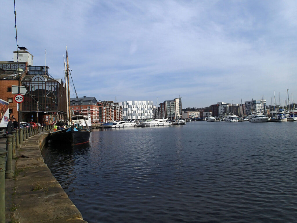 Modern Dockside buildings, one on stilts, sailing barge, water and modern cruisers, blue sky reflected