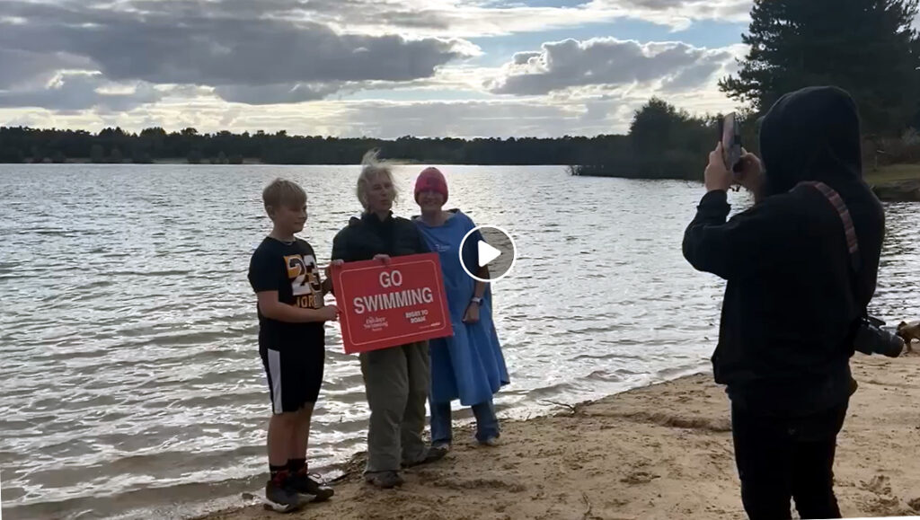 screenshot of video, three standing (one boy, two women) with filmer, in from of lake, with Go Swimming placard