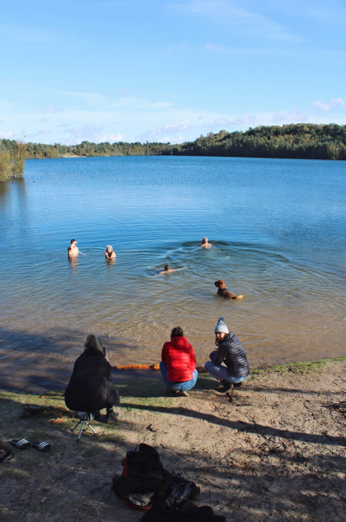 Swimmers and dog in large lake, water blue reflecting blue sky, people sitting on sandy beach