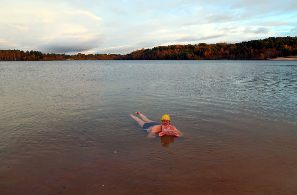 Swimmer lying in the shallow water with placard saying “Go Swimming” and a large lake behind her