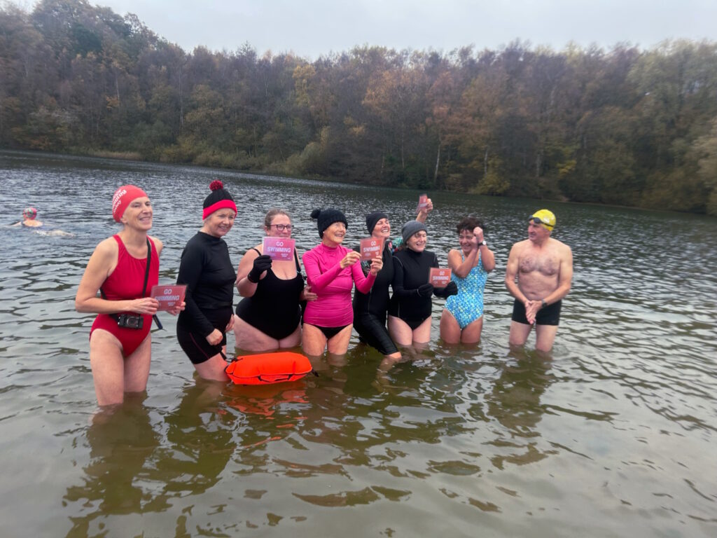 Swimmers standing in a lake in swim costumes, holding placards saying “Go Swimming”, sandy beach, wooded cliff behind them