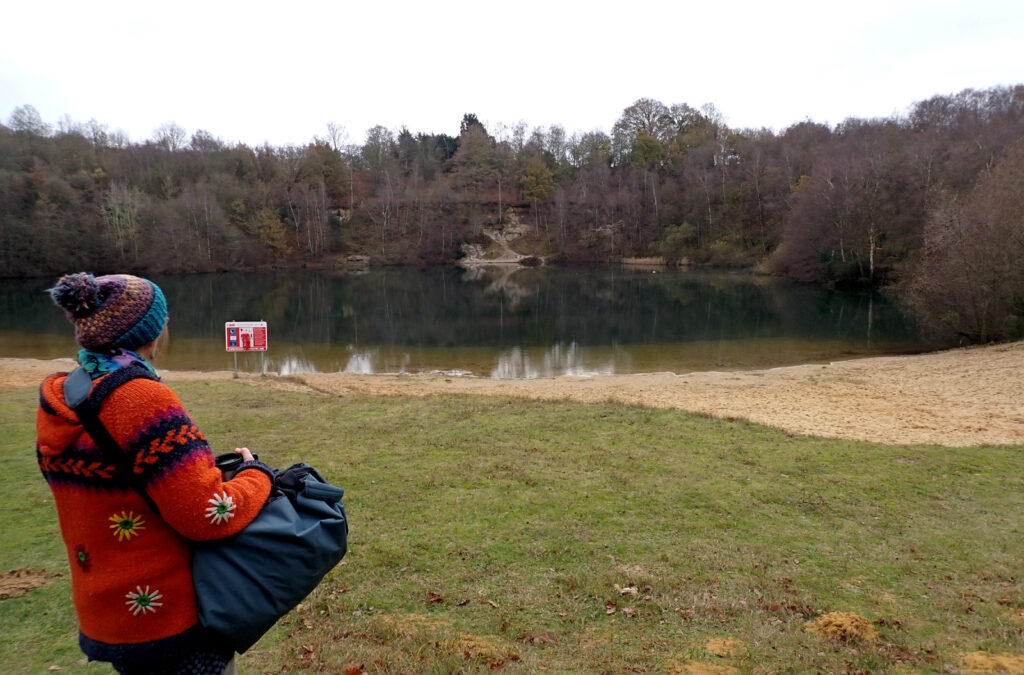 Woman in colourful cardigan and bobble hat with a view of a still lake, sandy beach and wooded backdrop, also a safety equipment station