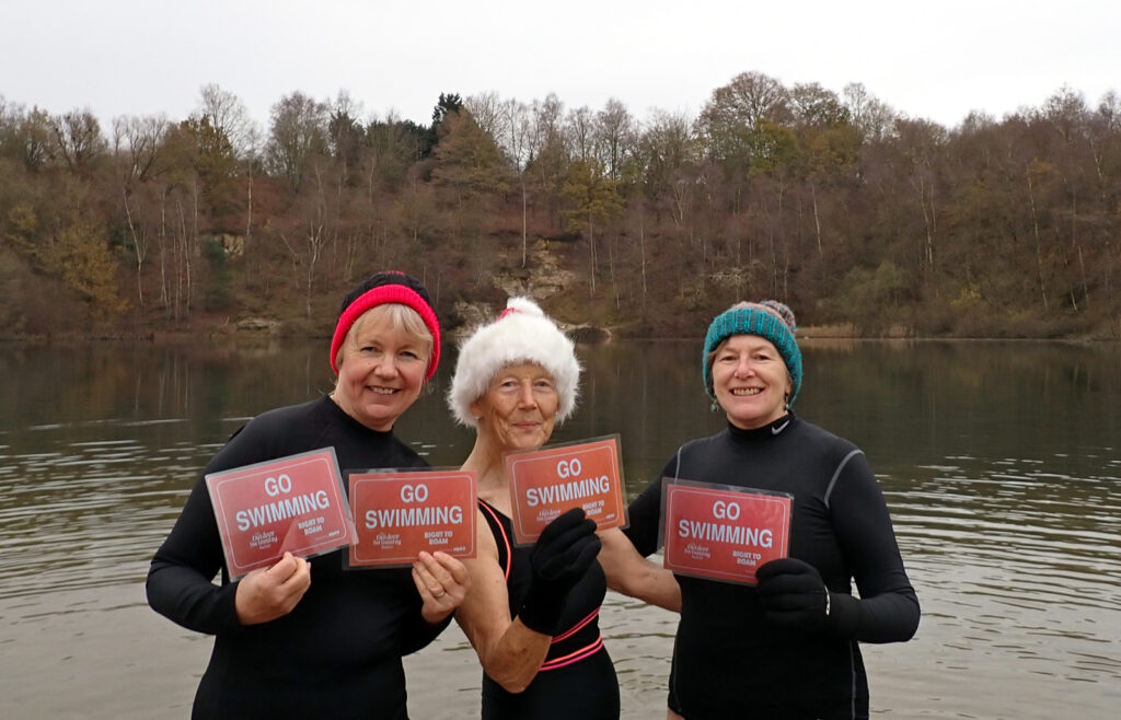 swimmers standing in a lake in swim costumes, holding placards saying “Go Swimming”, sandy beach, wooded cliff behind them