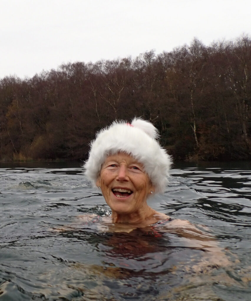 swimmer smiling in white Christmas hat in a lake with a wooded backdrop