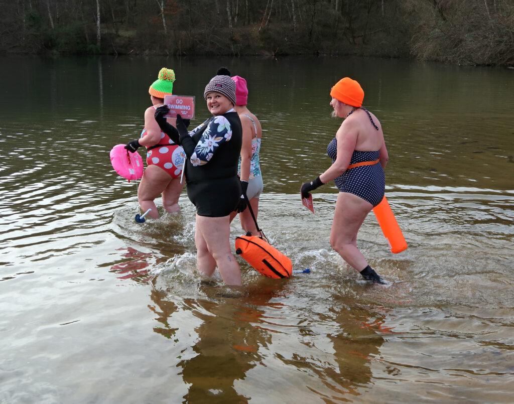 Four swimmers in swimming costumes in lake, one with a placard reading “Go Swimming”