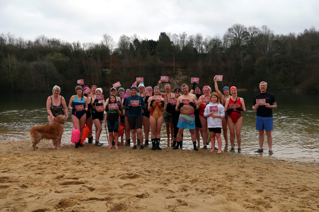 22 of the swimmers (and a dog) standing along the edge of the sandy beach holding placards reading “Go Swimming”