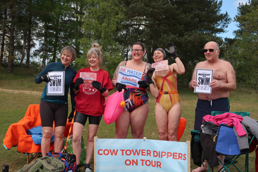 5 swimmers holding placards reading “Be Water Aware”, “Go Swimming” and “We Have the Right To Swim”, with banner reading “Cow Tower dippers on tour”