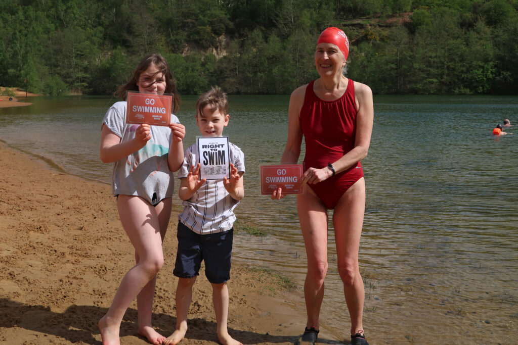 Two children and one swimmer standing along the edge of the sandy beach holding placards reading “Go Swimming” and “We Have the Right To Swim”